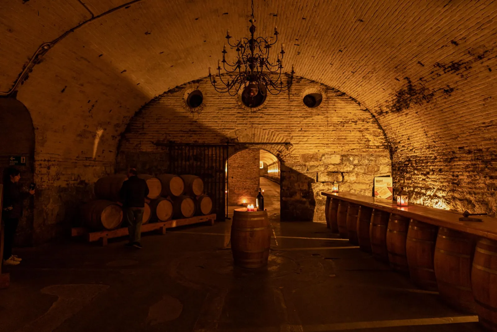 Candlelit vaulted brick cellar with a chandelier, oak barrels and a barrel tasting bar