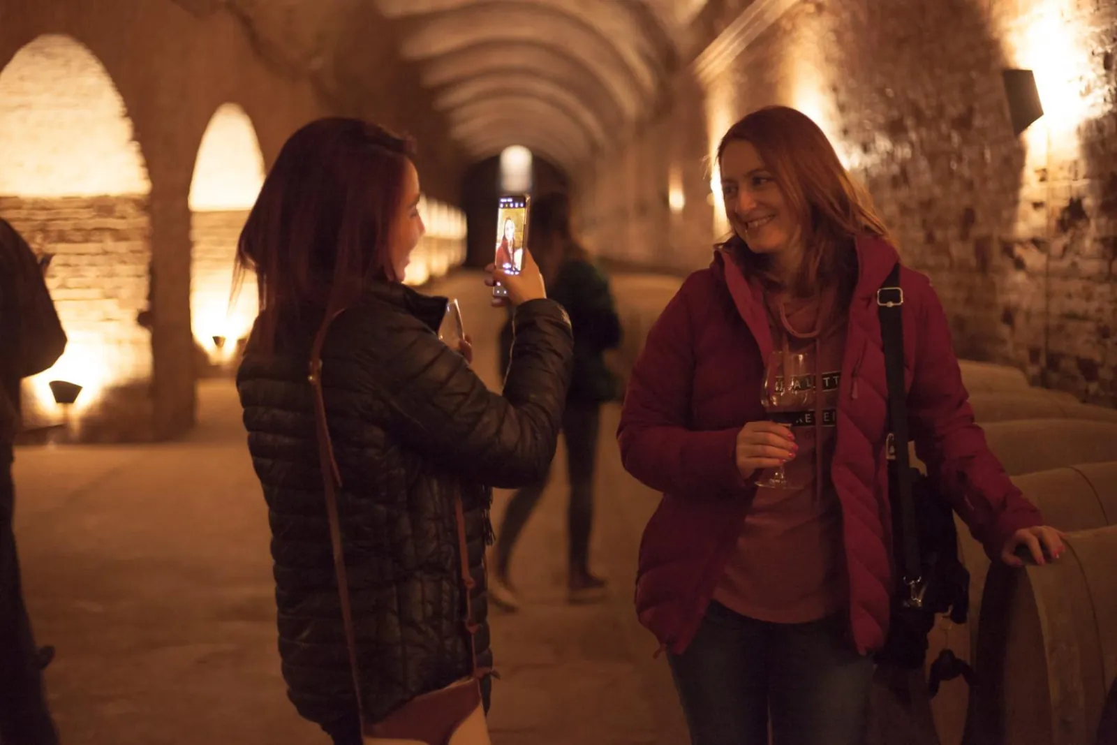 Visitor photographing a friend holding wine in an arched, lamp-lit cellar tunnel