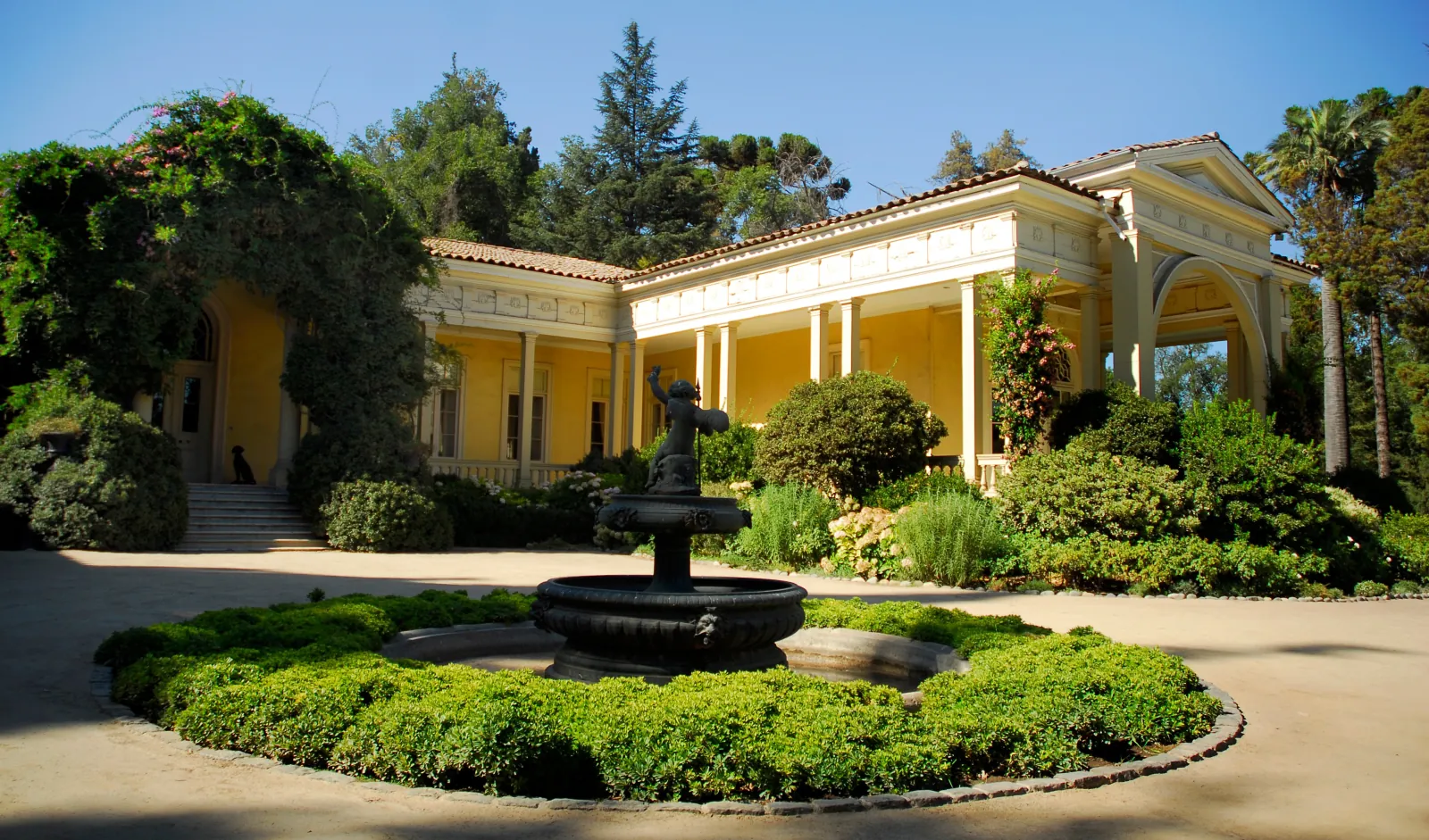 Yellow colonnaded house behind a fountain and clipped hedges in sunshine