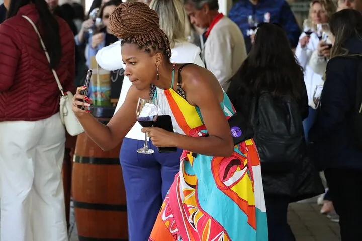 Woman in a colorful dress photographing her glass of red wine beside barrels