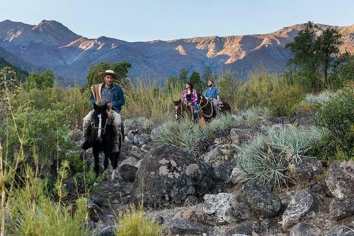 Huaso guide leading two riders along a rocky trail below mountains