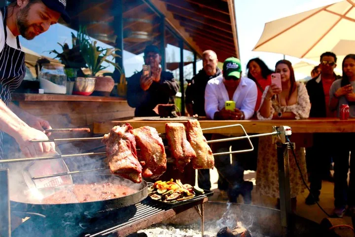 Chef grilling large cuts of meat over an open fire as diners watch